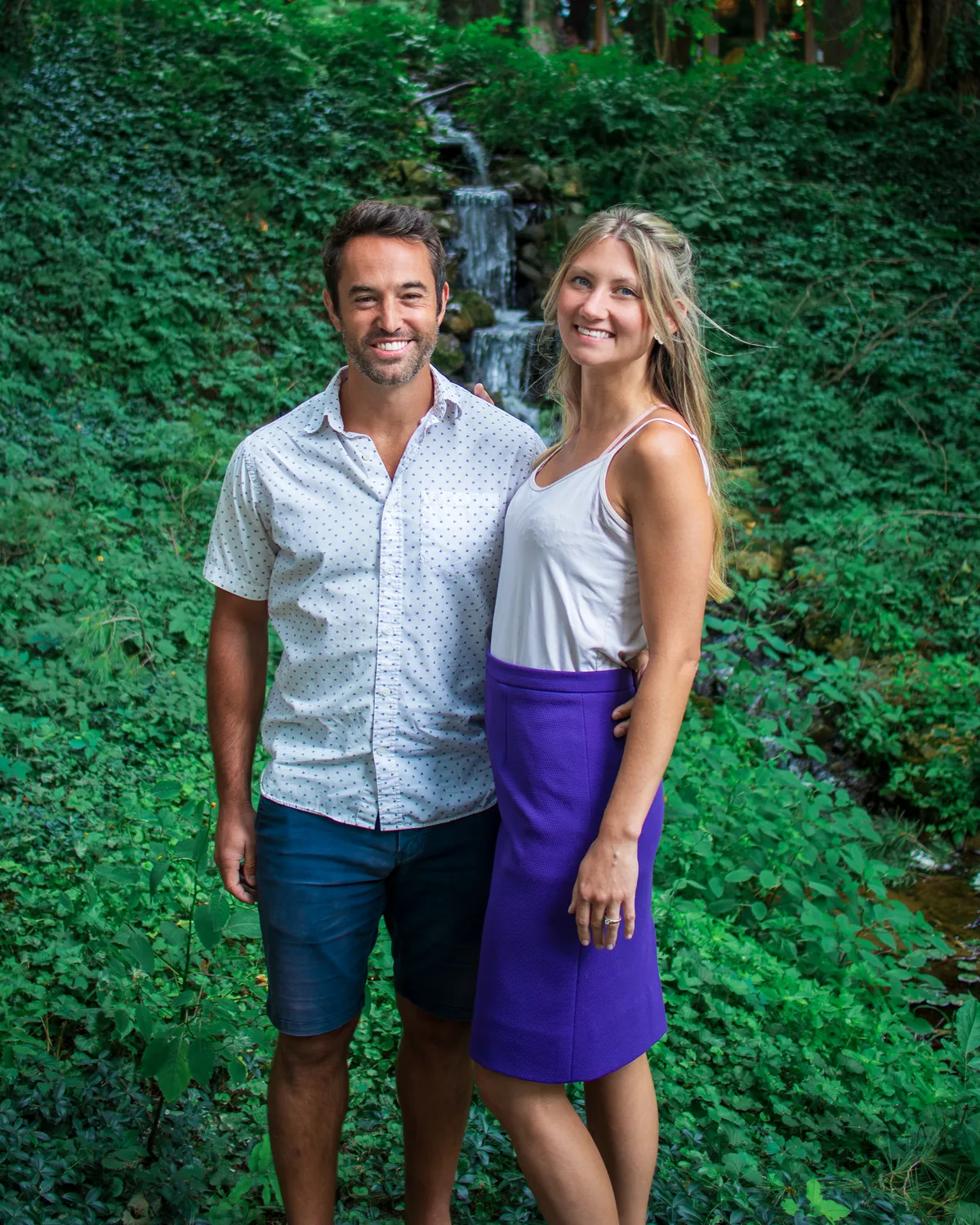 A man and a woman smiling together in front of a small waterfall in a green garden.